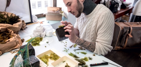 Le plumassier Maxime Leroy assis derrière son établis, en plein collage de plumes vertes 